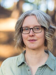 Picture of Sally Moss, with curled hair and light green open-necked shirt, standing smiling against a backdrop of blurry trees